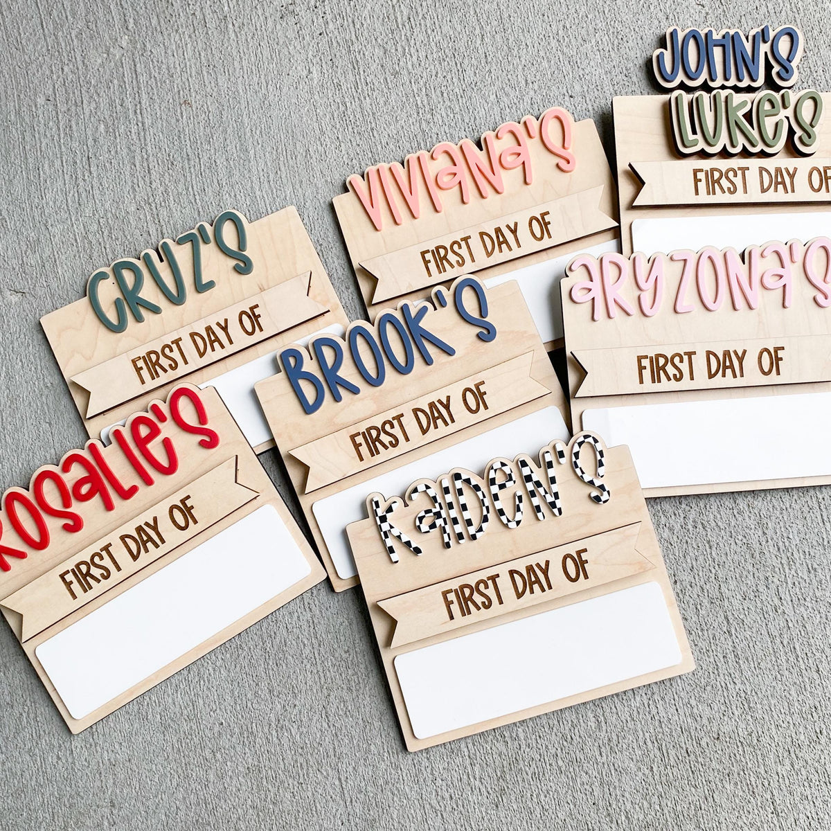 Flat lay of many personalized simple back to school boards spread on a gray wood surface, each engraved with a first day of header and a different child's name such as Cruz's, Rosalie's, Vivian's, Brooks, and Grayson's in assorted bubble letter colors, with blank dry erase strips.