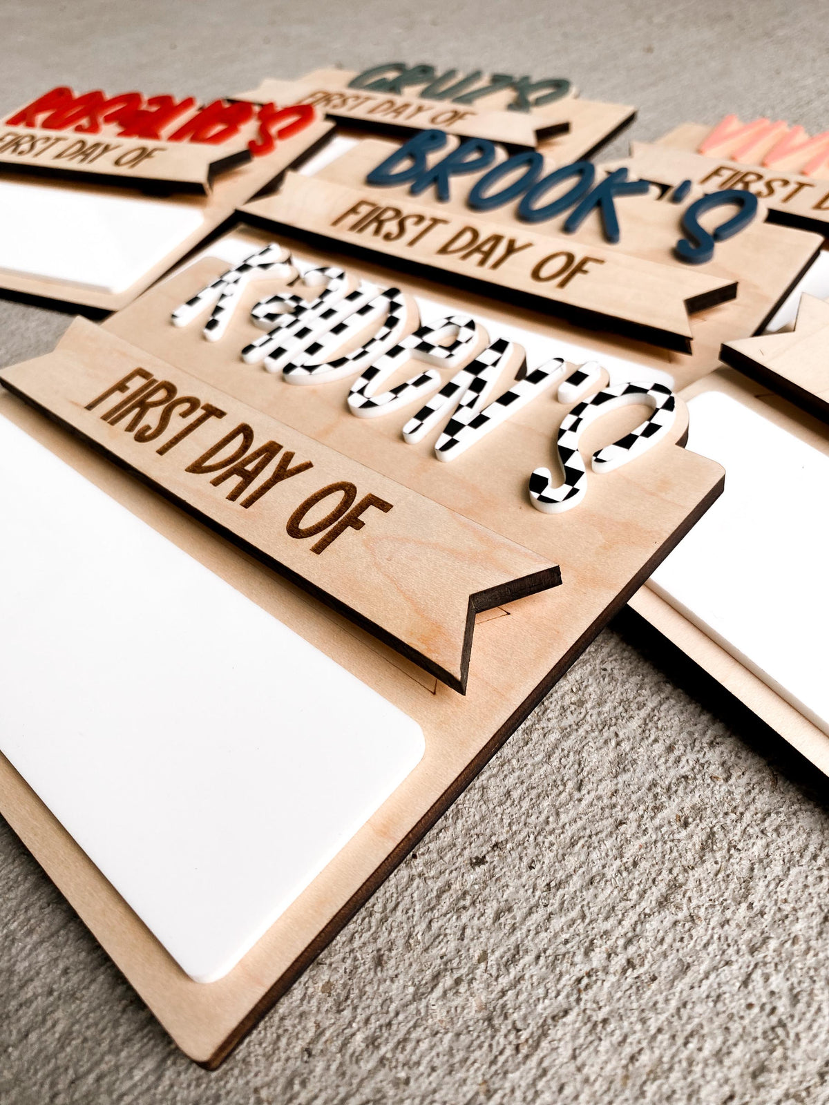 A collection of personalized simple back to school boards stacked at an angle, showing names like Maddox, Brooks, and Kennedy in red, blue, and spotted black-and-white lettering, each with a first day of header and blank dry erase strip, demonstrating custom name and color choices.