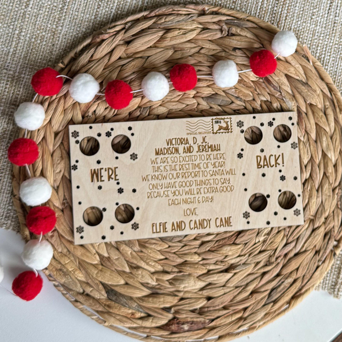 Two elf We're Back postcard on a round woven placemat with a red and white pom pom garland, ready for elf arrival day