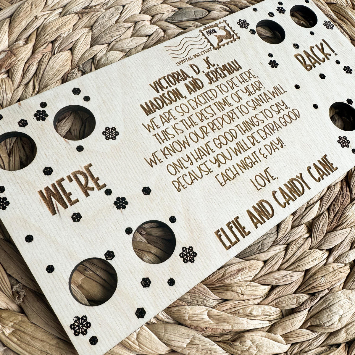 Angled close-up of the two elf We're Back message engraved in maple wood with snowflake details and personalized names