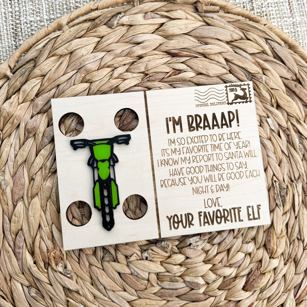Dirt bike elf welcome back postcard in maple wood with a green dirt bike and I'm Braaap message, on a woven placemat