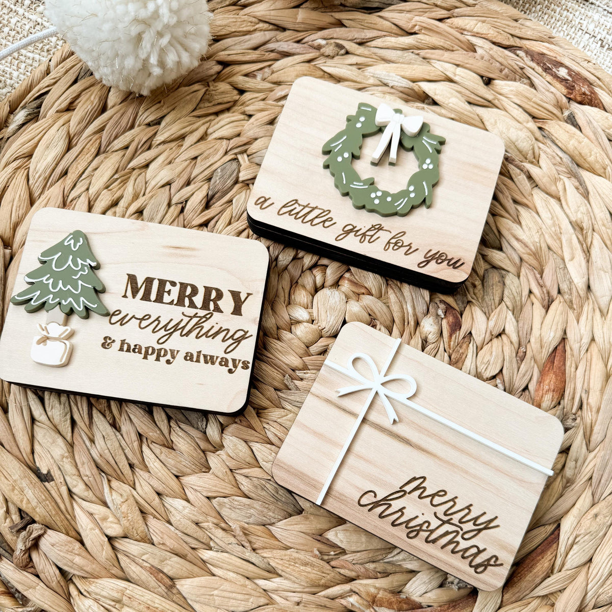 Close-up of three wooden Christmas money holders with raised acrylic wreath, tree, and white bow details and engraved holiday greetings
