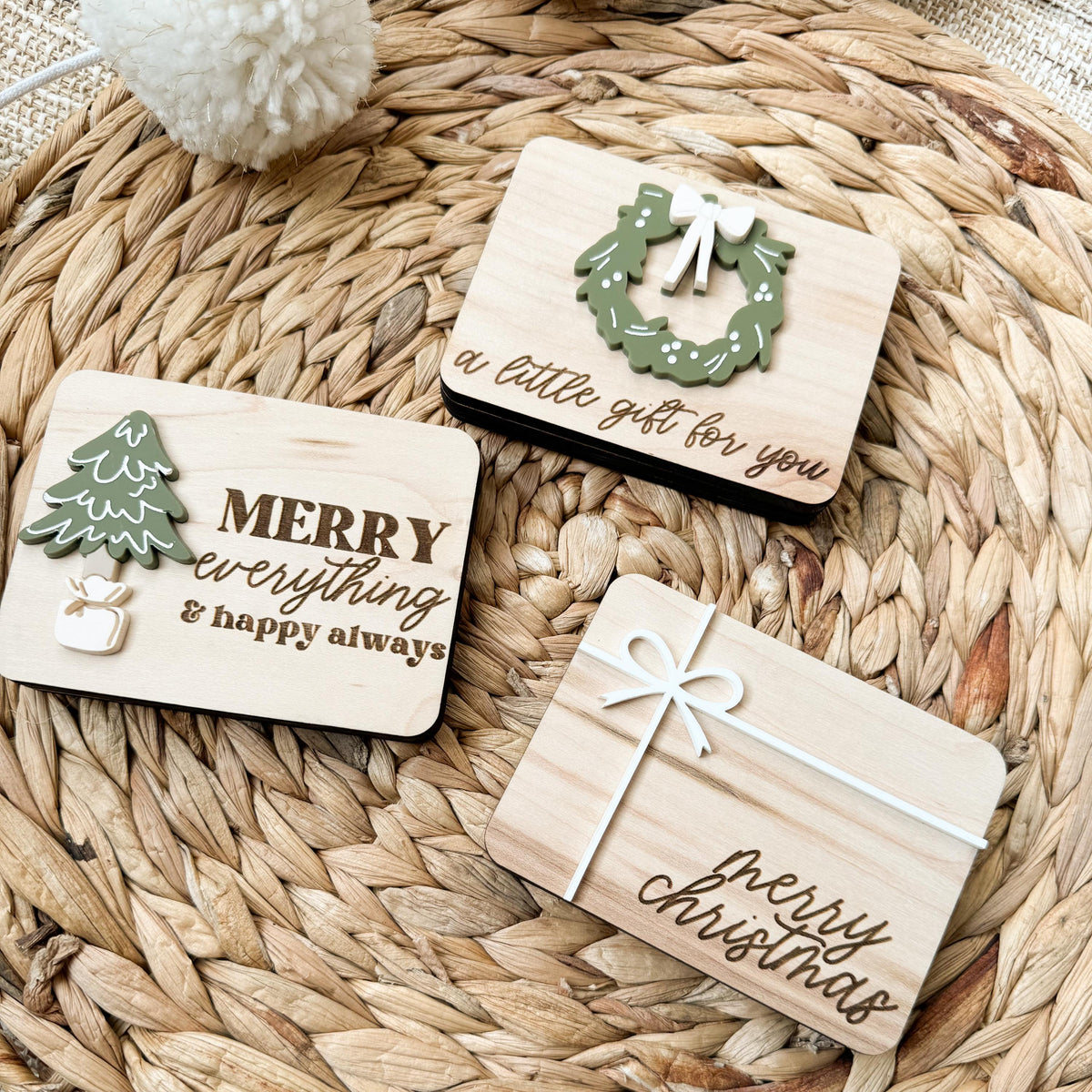 Close-up of three Christmas gift card holders made of maple wood with raised acrylic wreath, tree, and white bow details and engraved holiday greetings
