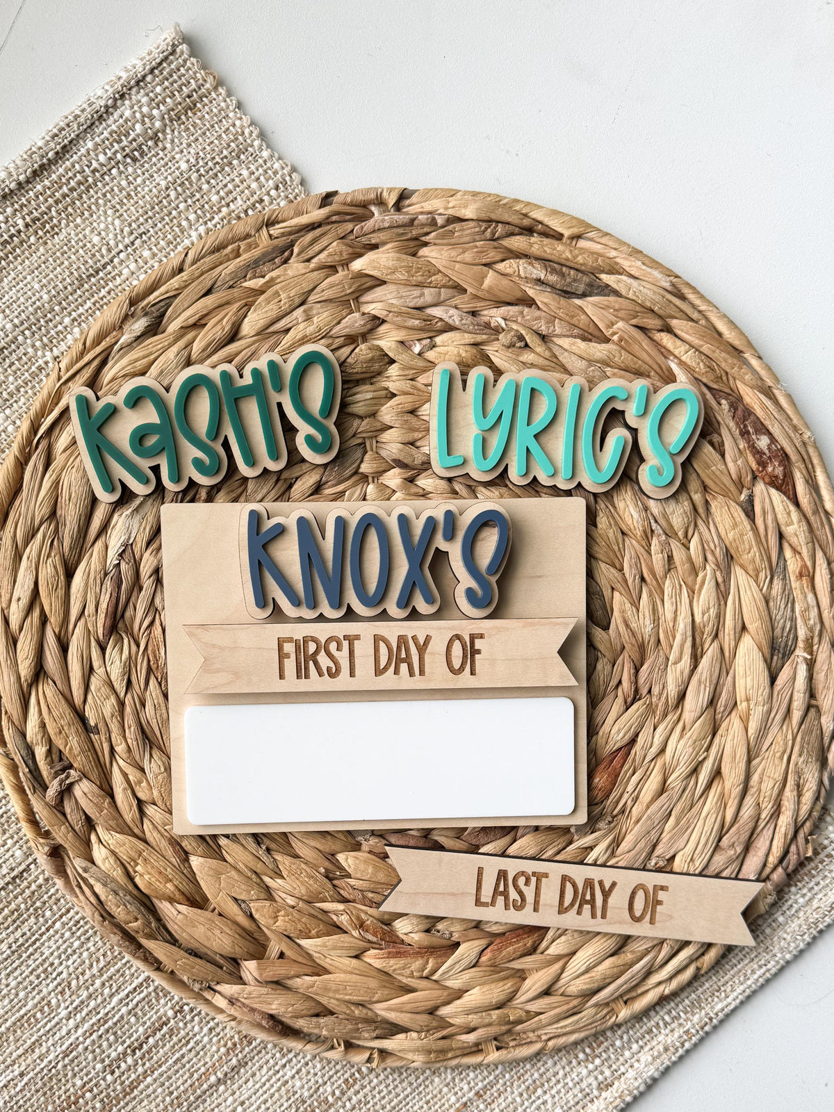 Personalized name pieces in teal and blue bubble letters reading Kash's, Lyric's, and Knox's shown with a back to school board's first day of and last day of interchangeable header strips and a blank dry erase strip, arranged on a round woven placemat to show custom name color options.