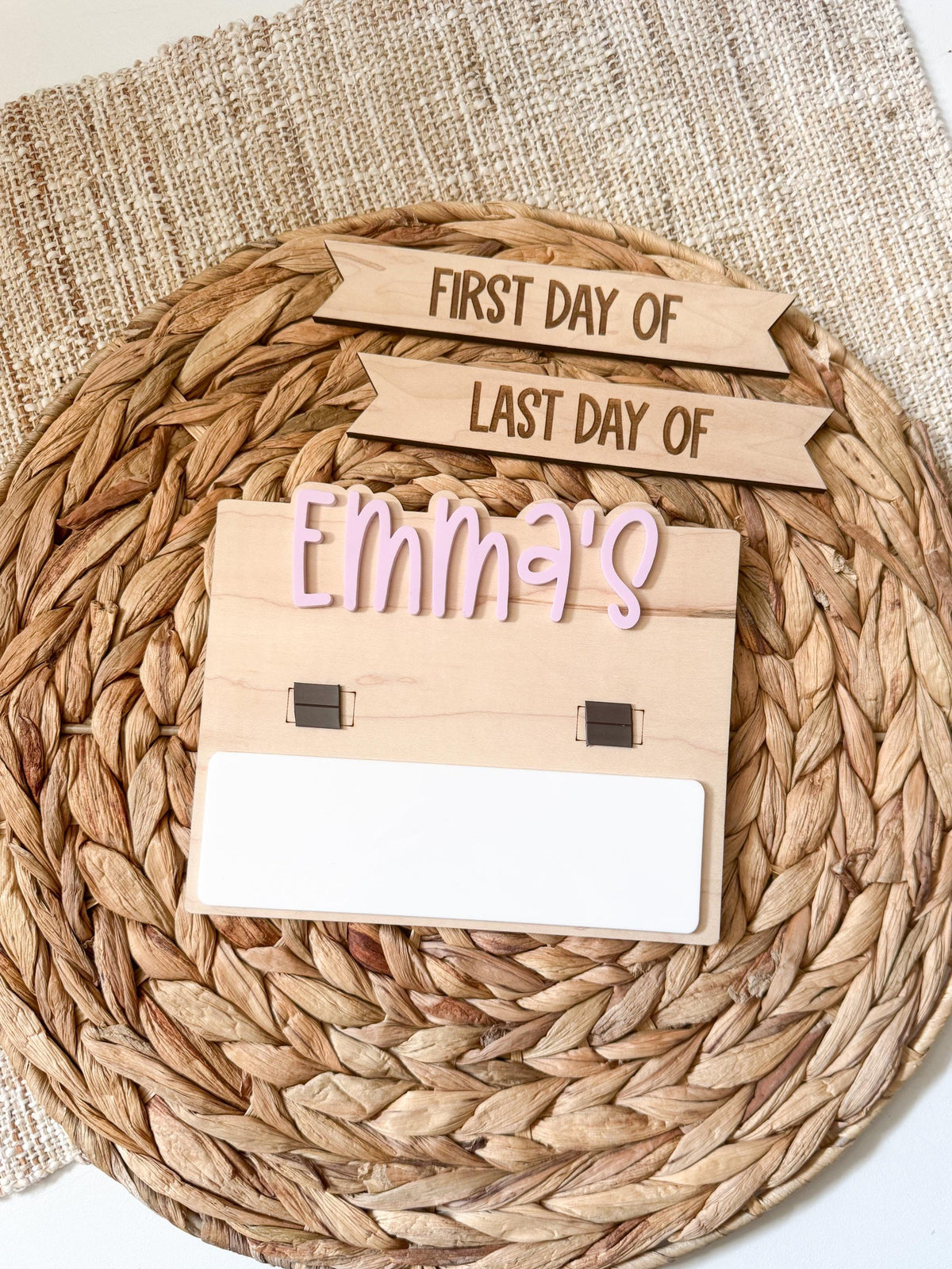 aPersonalized simple back to school board with the name Emma's in purple bubble letters, displayed with both first day of and last day of interchangeable header strips above the black magnetic tabs and a single blank dry erase strip, on a round woven placemat.