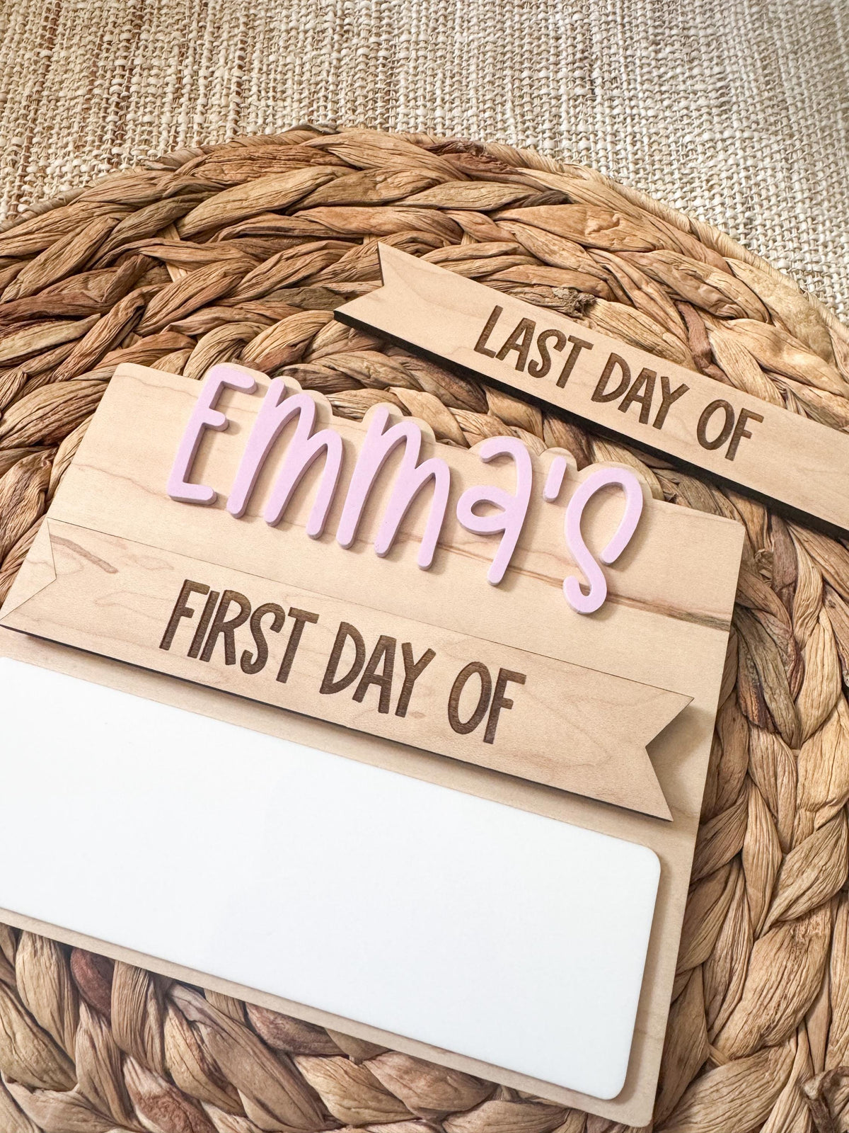 Angled close-up of the personalized Emma's simple back to school board in soft purple bubble letters, with the interchangeable last day of strip and first day of header above a blank white dry erase strip, on a round woven placemat.
