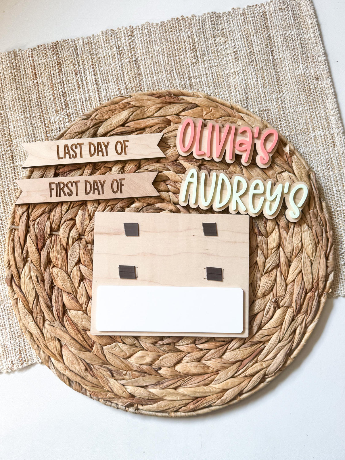 Personalized simple back to school board on a round woven placemat showing the black magnetic name tabs, with Olivia's and Audrey's name pieces in pink and cream above interchangeable last day of and first day of header strips and a blank dry erase strip.