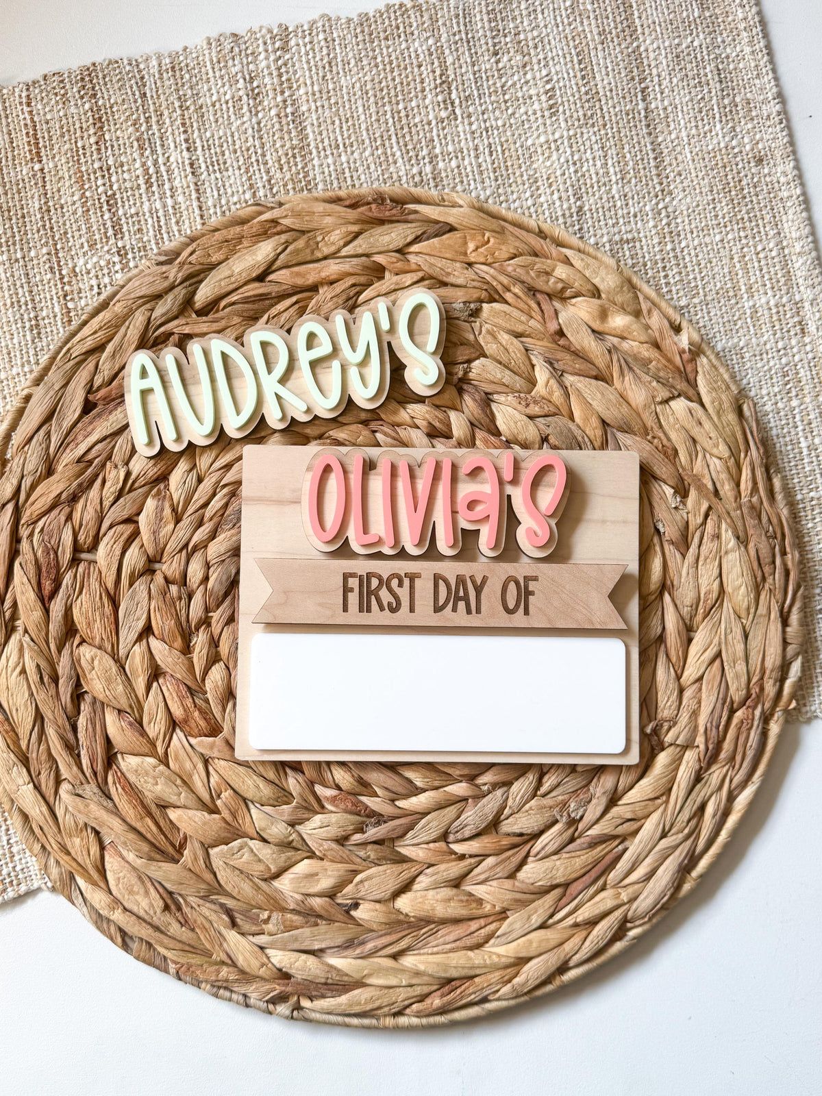 Personalized simple back to school board on a round woven placemat with Olivia's installed in pink bubble letters above a first day of header and blank dry erase strip, and an Audrey's name piece in cream shown above as an interchangeable name option.