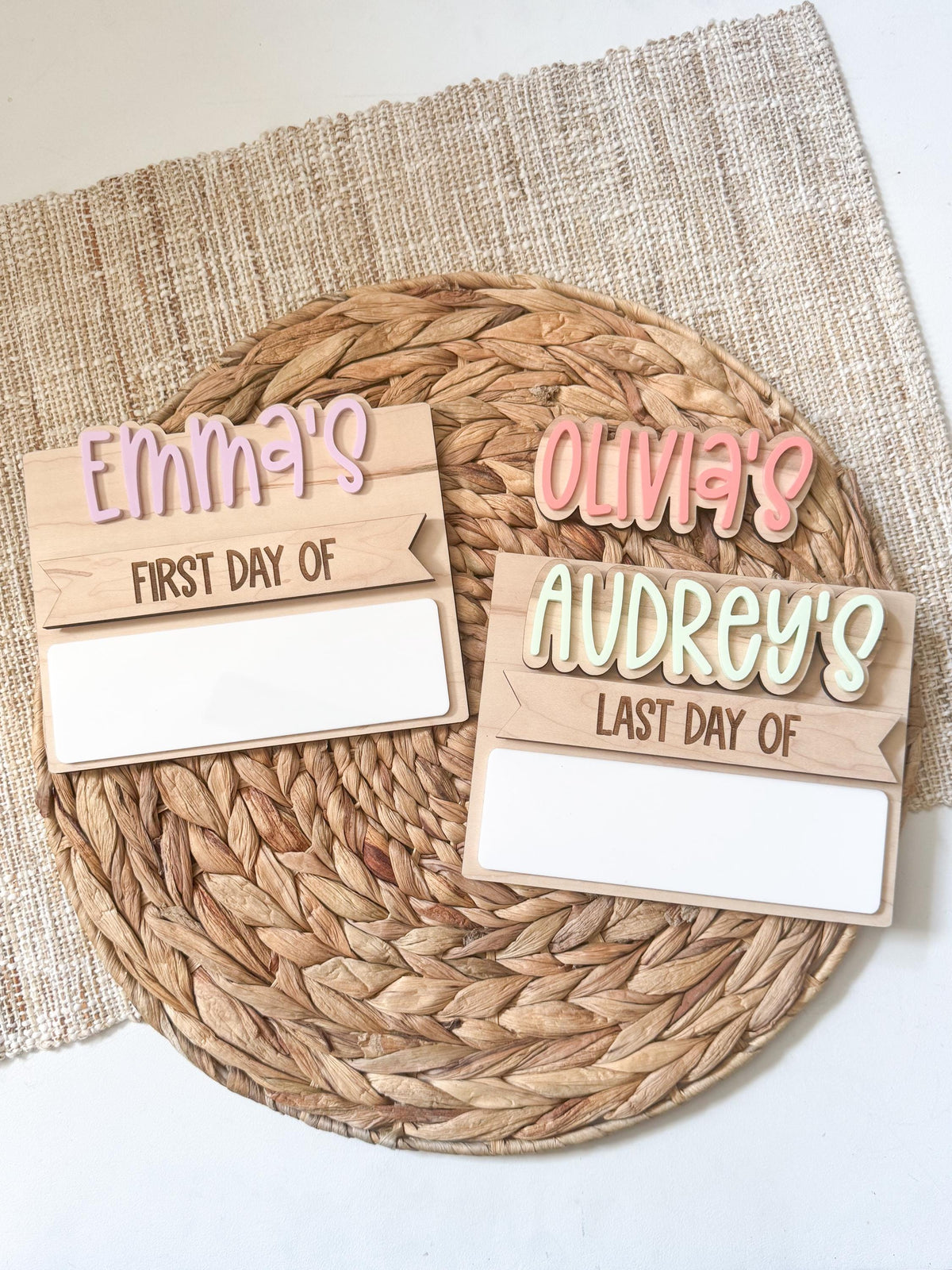 Two personalized simple back to school boards on a woven placemat, one with Emma's in purple bubble letters and a first day of label, the other showing Olivia's and Audrey's name pieces in pink and cream with a last day of strip, each with a blank dry erase field for a reusable school photo prop.