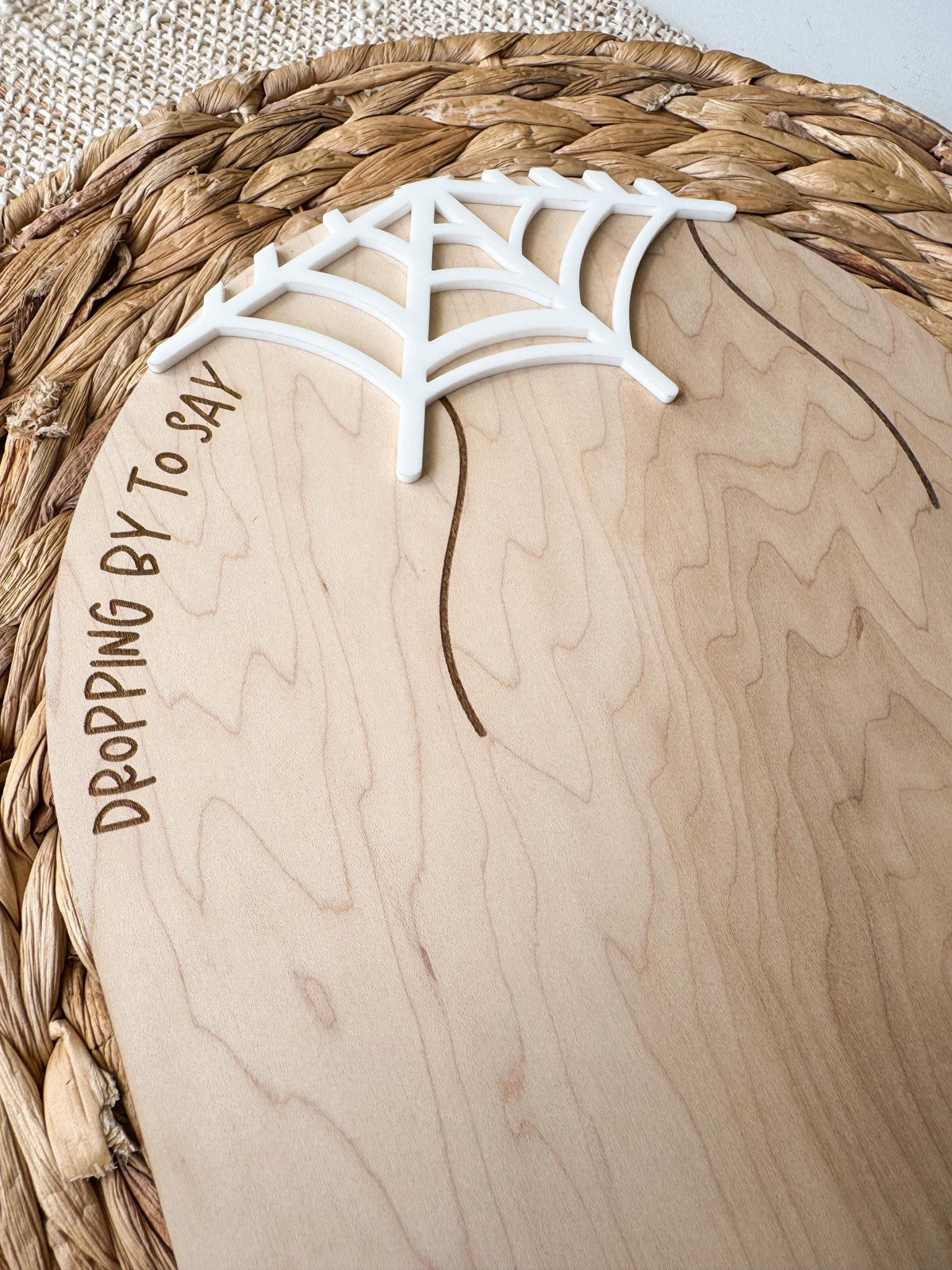 Close-up of the raised white acrylic spider web on the curved top of the maple wood board. Engraved “Dropping by to say” lettering and two hanging lines sit against the visible natural wood grain.