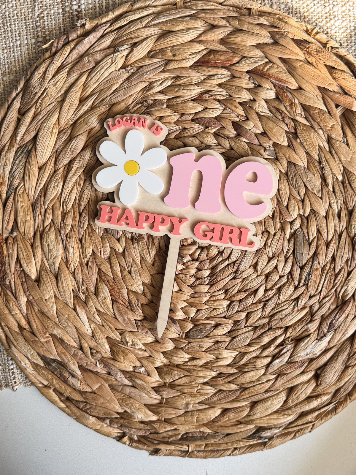 Acrylic One Happy Girl first birthday cake topper with white daisy and personalized name, top-down view on wicker charger with white surface
