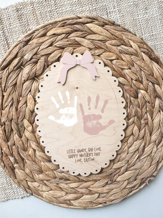 Personalized maple wood handprint sign with scalloped edge and pink acrylic bow, featuring two painted handprints and custom Mother’s Day message, displayed on a woven rattan background.