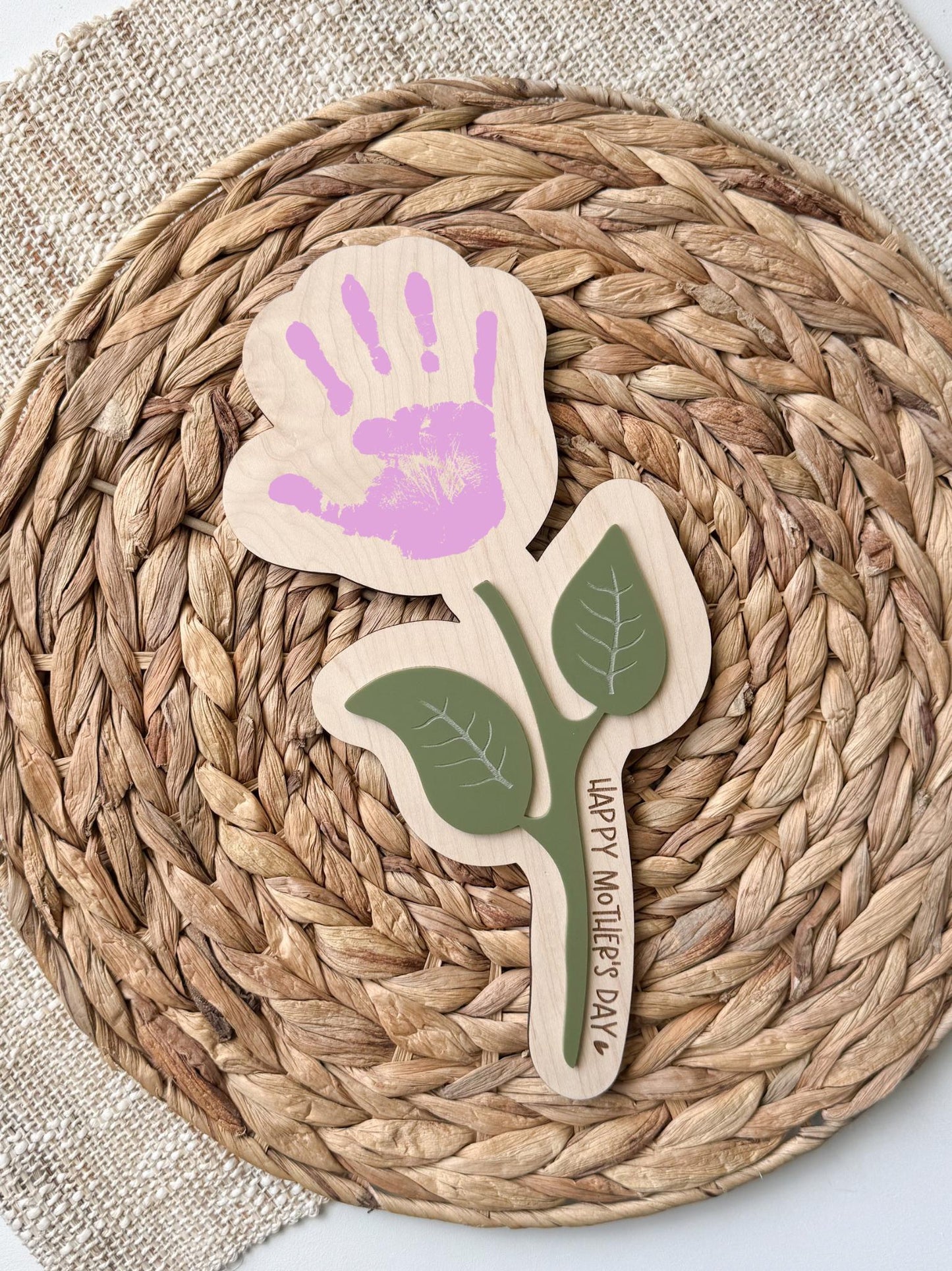 Personalized maple wood handprint flower sign with sage green acrylic leaf accents and custom “Happy Mother’s Day” message along the stem, displayed on a woven rattan background.