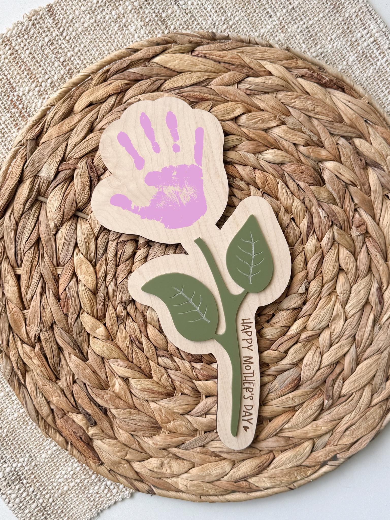 Personalized maple wood handprint flower sign with sage green acrylic leaf accents and custom “Happy Mother’s Day” message along the stem, displayed on a woven rattan background.