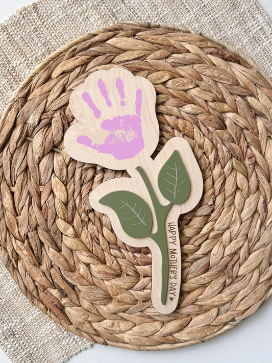 Personalized maple wood handprint flower sign with sage green acrylic leaf accents and custom “Happy Mother’s Day” message along the stem, displayed on a woven rattan background.
