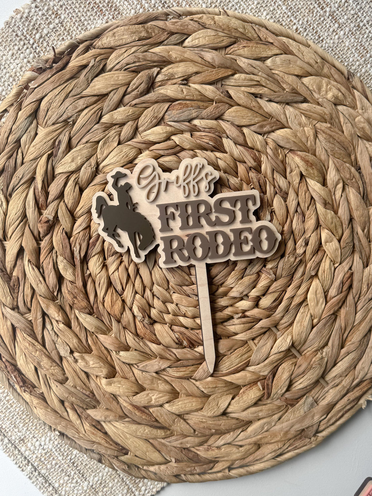 Neutral-toned first rodeo cake topper with layered acrylic lettering, personalized name, cowboy bronc rider silhouette, and natural maple wood backing.
