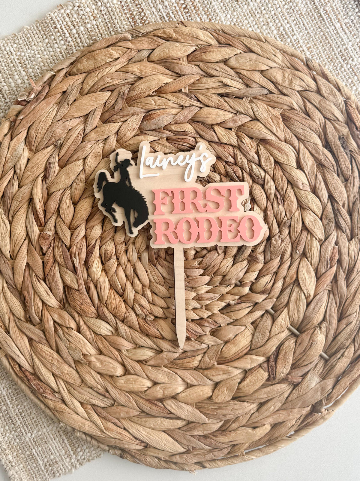 Personalized first rodeo cake topper with a black cowgirl bronc rider silhouette, layered pink acrylic lettering, custom script name, and natural wood backing displayed on a woven mat.
