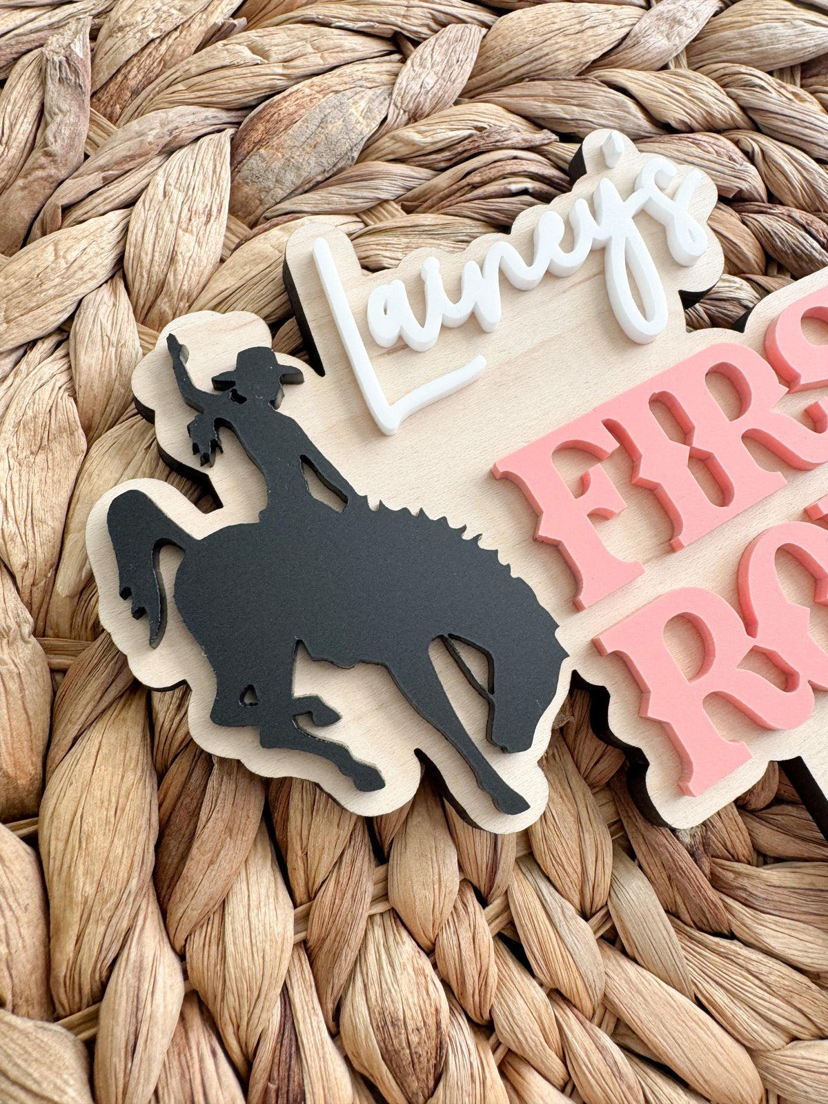 Close-up of the layered acrylic name and cowgirl bronc rider silhouette on a personalized western birthday cake topper with natural wood grain.
