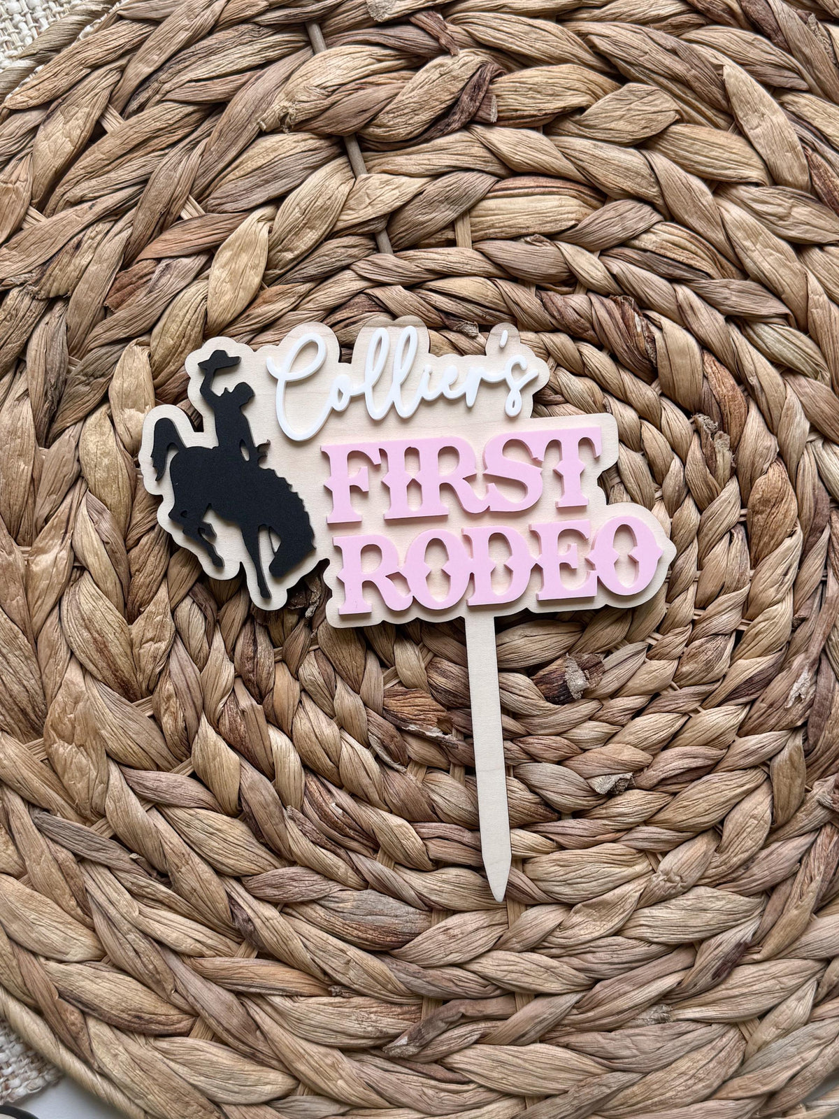 Wooden first rodeo cake topper with layered acrylic name, pink birthday lettering, and cowgirl bronc rider silhouette for a western birthday celebration.