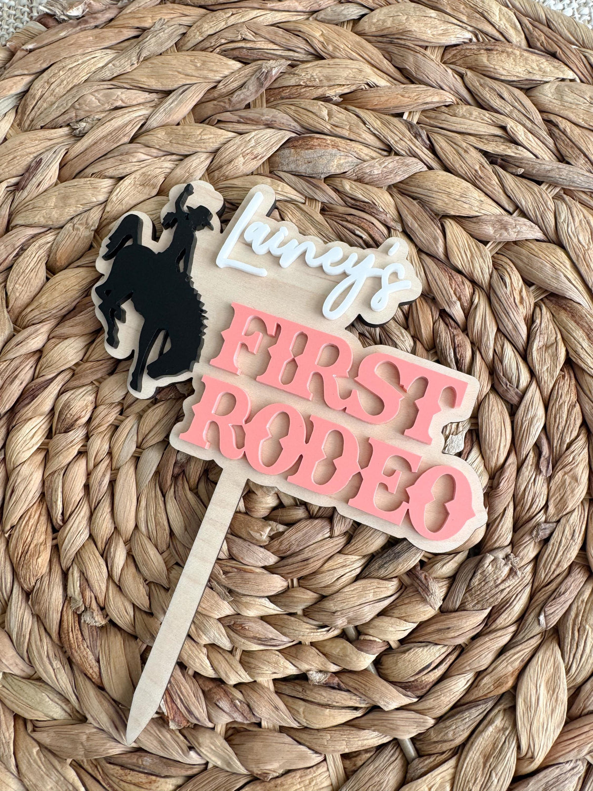 Personalized first rodeo cake topper with a black cowgirl bronc rider silhouette, layered pink acrylic lettering, custom script name, and natural wood backing displayed on a woven mat.