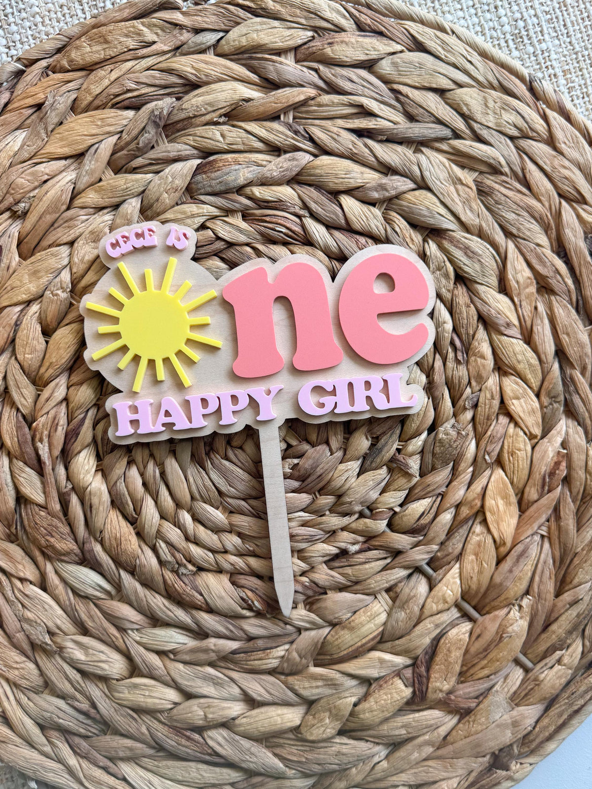 Personalized One Happy Girl sunshine cake topper displayed on woven background for a girl’s first birthday.