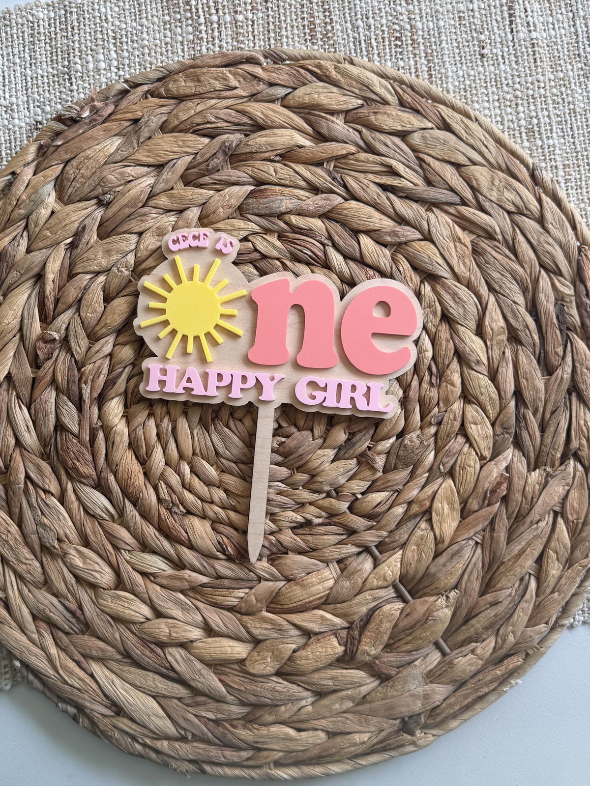 Personalized sunshine cake topper with custom name shown on woven mat for first birthday party decor.