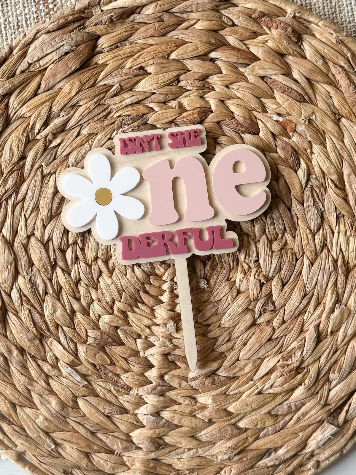 Personalized daisy One-derful cake topper displayed on woven backdrop for first birthday party decor.