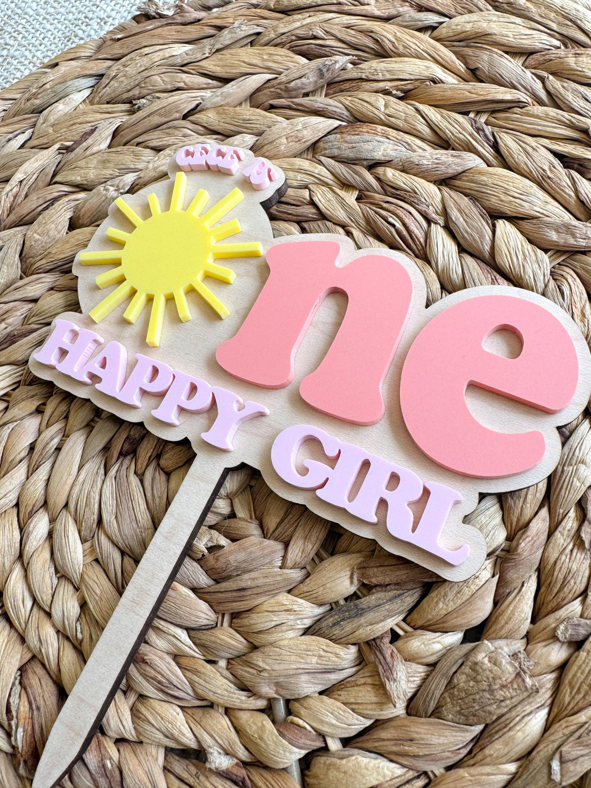 Angled view of custom sunshine birthday cake topper with layered maple wood and pink acrylic lettering.