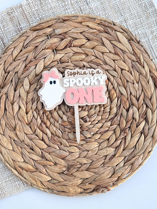 Personalized Spooky One first birthday cake topper with a white ghost wearing a pink bow and the child's name above SPOOKY ONE lettering in layered wood and acrylic, on a woven placemat.