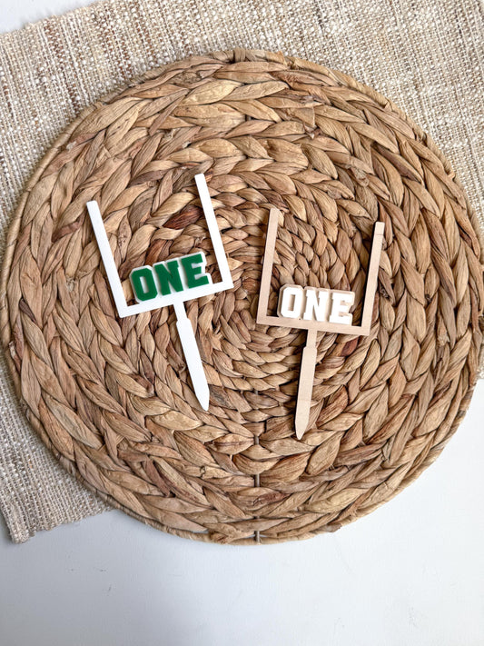 Two football goal post first birthday cake toppers with the word ONE, one in white with green lettering and one in natural wood with white lettering, displayed on a woven placemat.