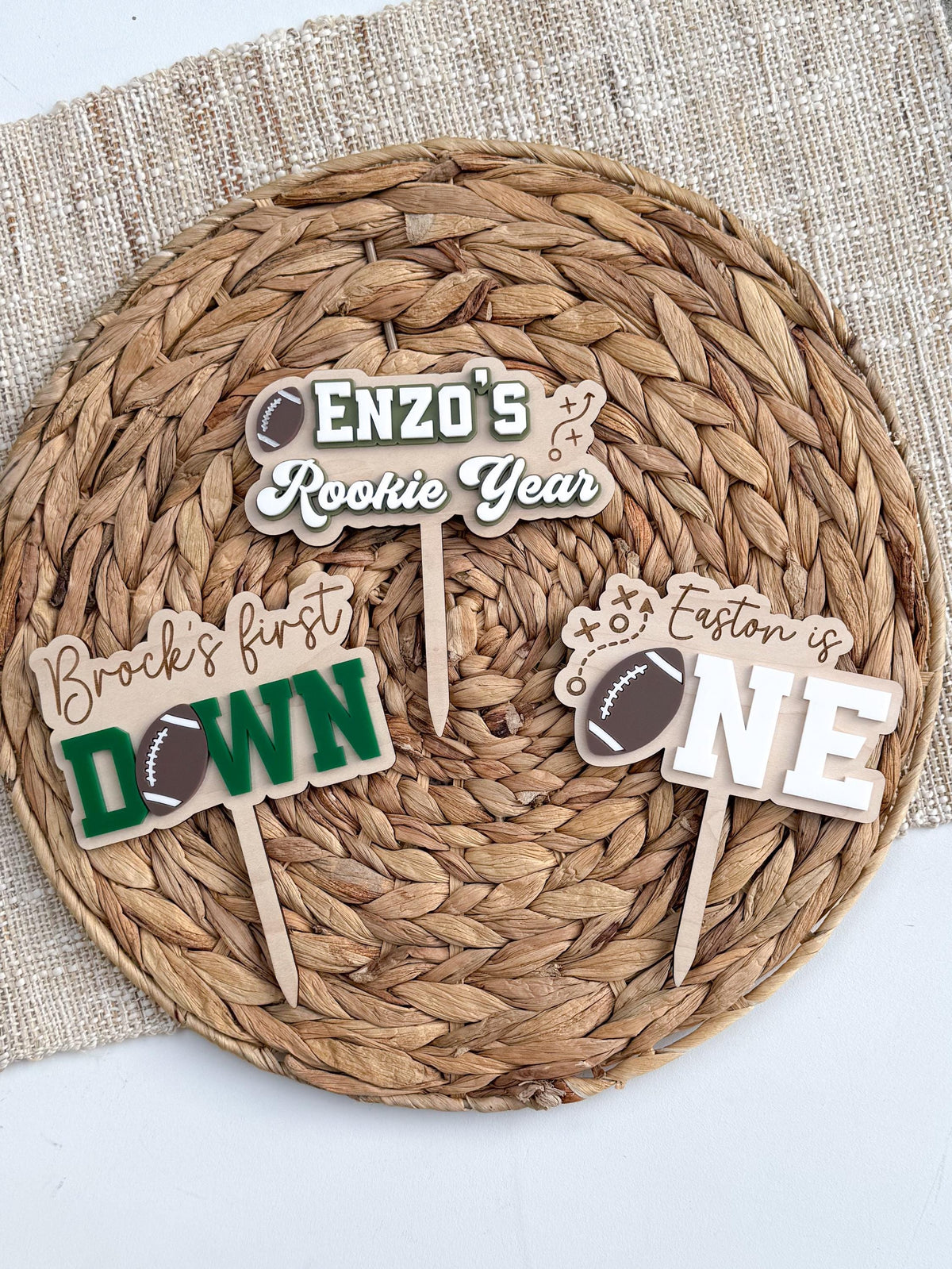 Three personalized football birthday cake toppers on a woven placemat: a Rookie Year design, a green and white First Down design and the brown and white football ONE design.