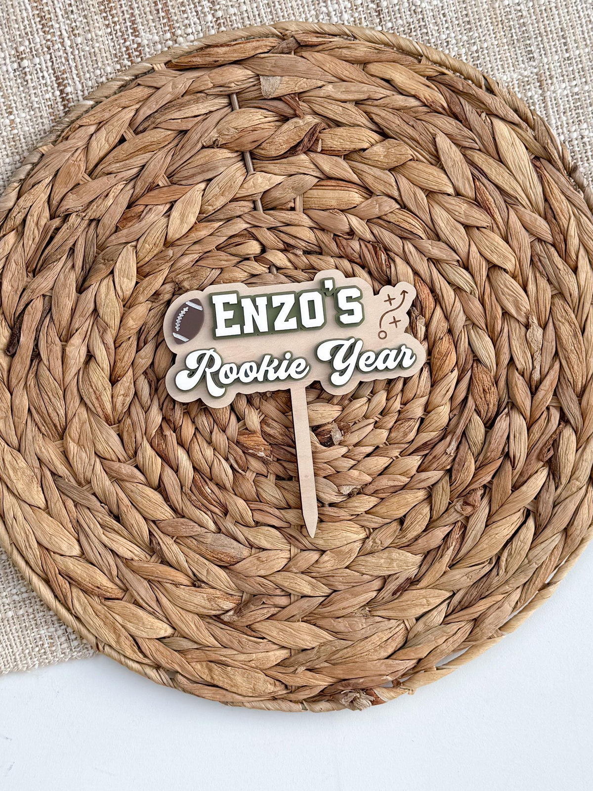 Overhead view of Rookie Year football cake topper on a round woven charger and linen, showing the green, white and natural wood colors and attached cake stake.