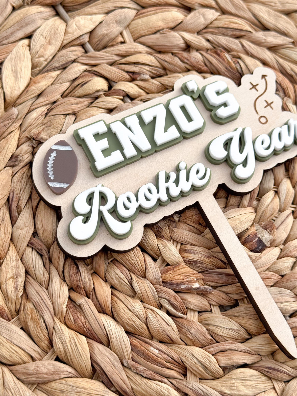 Angled close-up of Rookie Year cake topper showing raised white and green acrylic name and script lettering, layered brown football and maple wood backer.