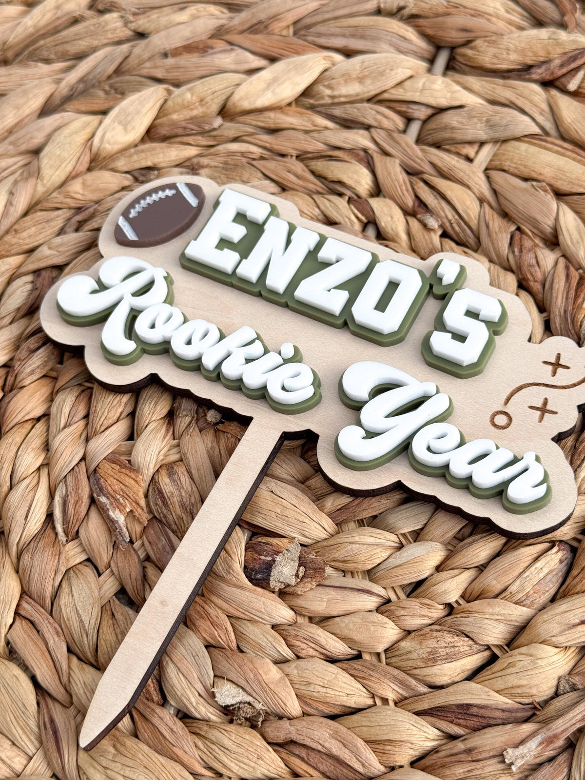 Tilted view of personalized Rookie Year football cake topper showing layered acrylic depth, engraved football play detail and attached maple wood cake stake.