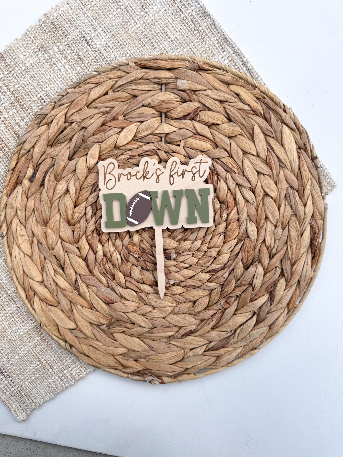 Sage green First Down football cake topper with engraved name and attached wood cake stake, displayed on a woven placemat for a football first birthday party
