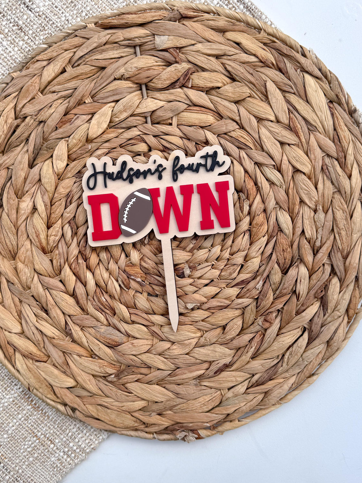 Red Fourth Down cake topper on a natural wood backer with engraved script name and attached cake stake, displayed on a woven placemat