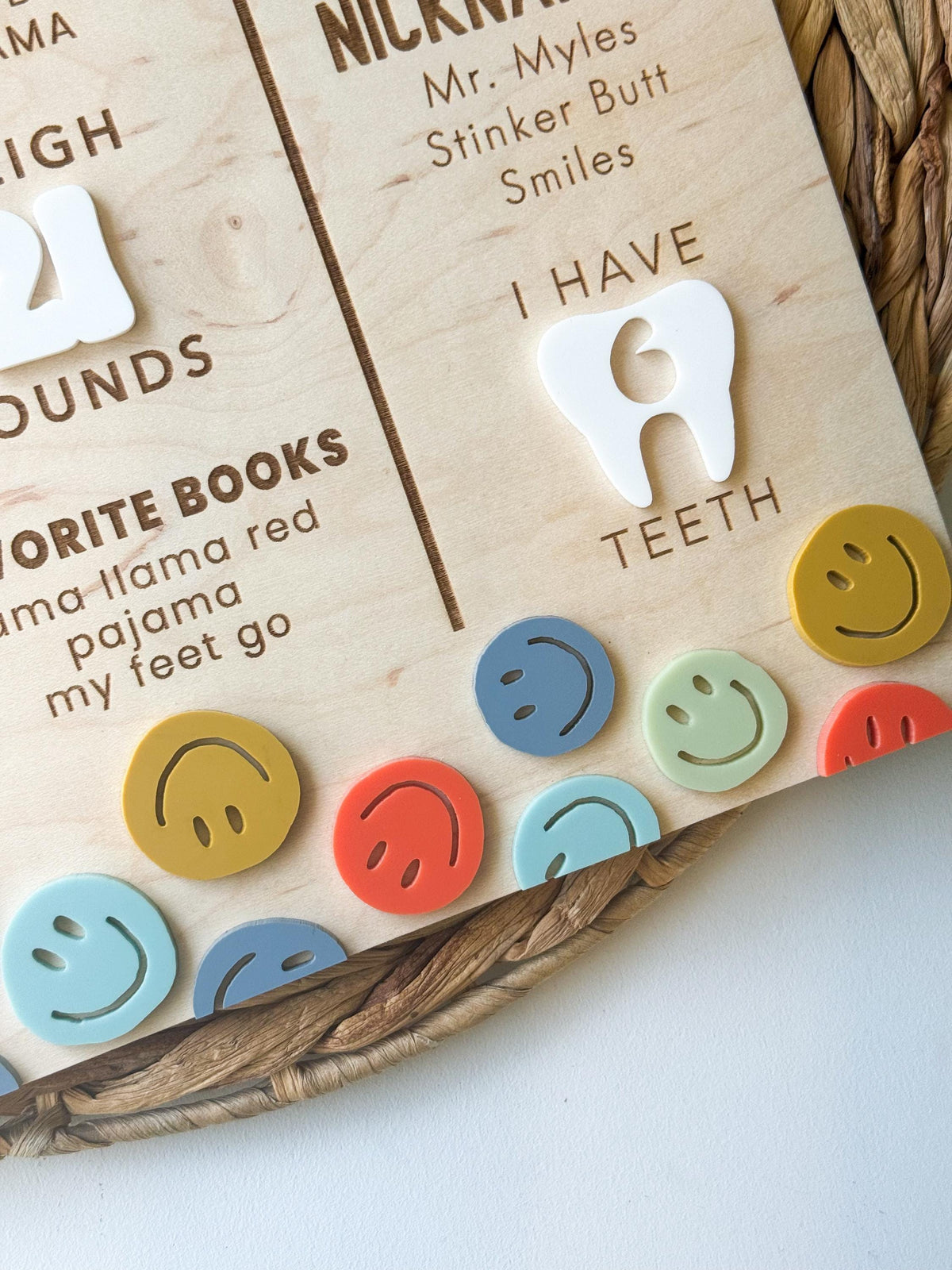 Angled close-up of colorful acrylic smiley face accents, a white acrylic tooth and engraved favorite books on a first birthday milestone board