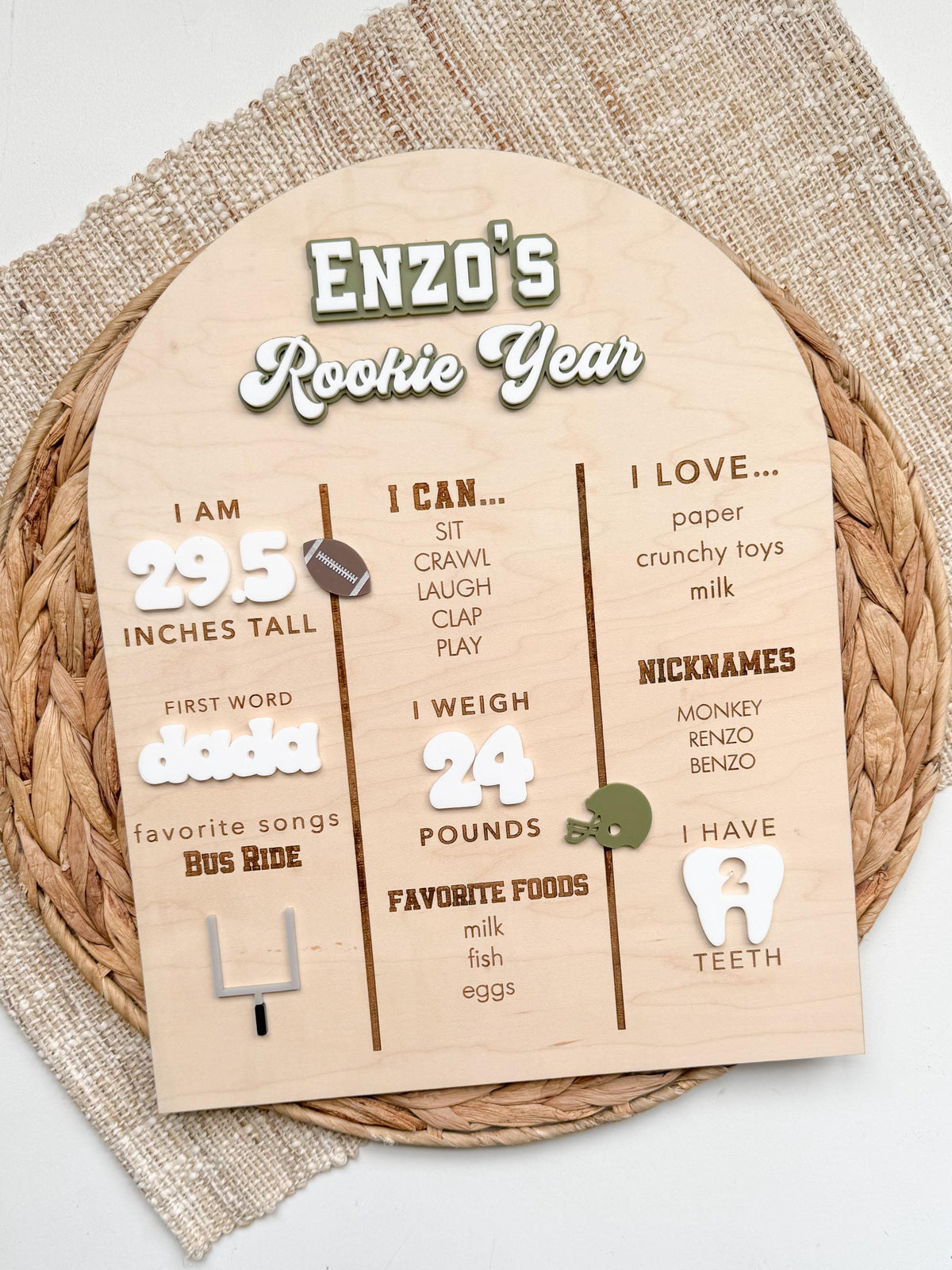 Football first birthday stat board in natural maple wood with a custom name and Rookie Year title in layered acrylic, engraved I am, I can and I love sections, white acrylic milestone numbers and football themed accents, styled on a woven placemat for first birthday party decor