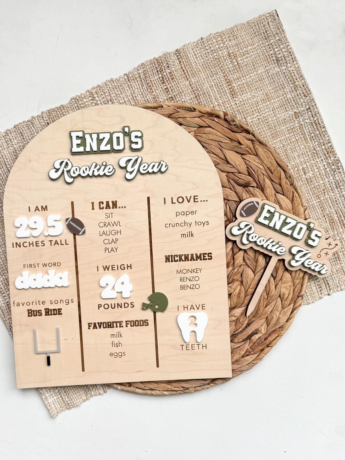 Rookie Year football first birthday milestone board shown with a coordinating personalized Rookie Year acrylic cake topper in matching green and white, a set of first birthday party decor styled on a woven placemat