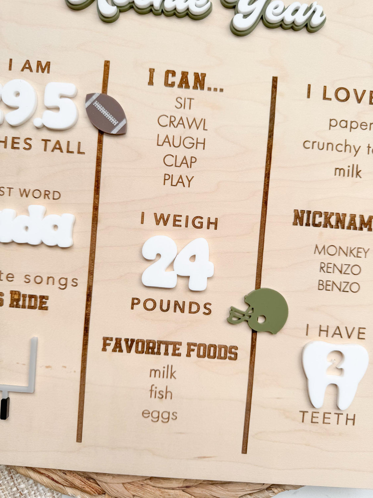 Detail of a personalized baby milestone sign showing engraved I can skills, white acrylic weight numbers, an olive green acrylic football helmet, engraved favorite foods, nicknames and I love lists, and a white acrylic tooth showing number of teeth on maple wood