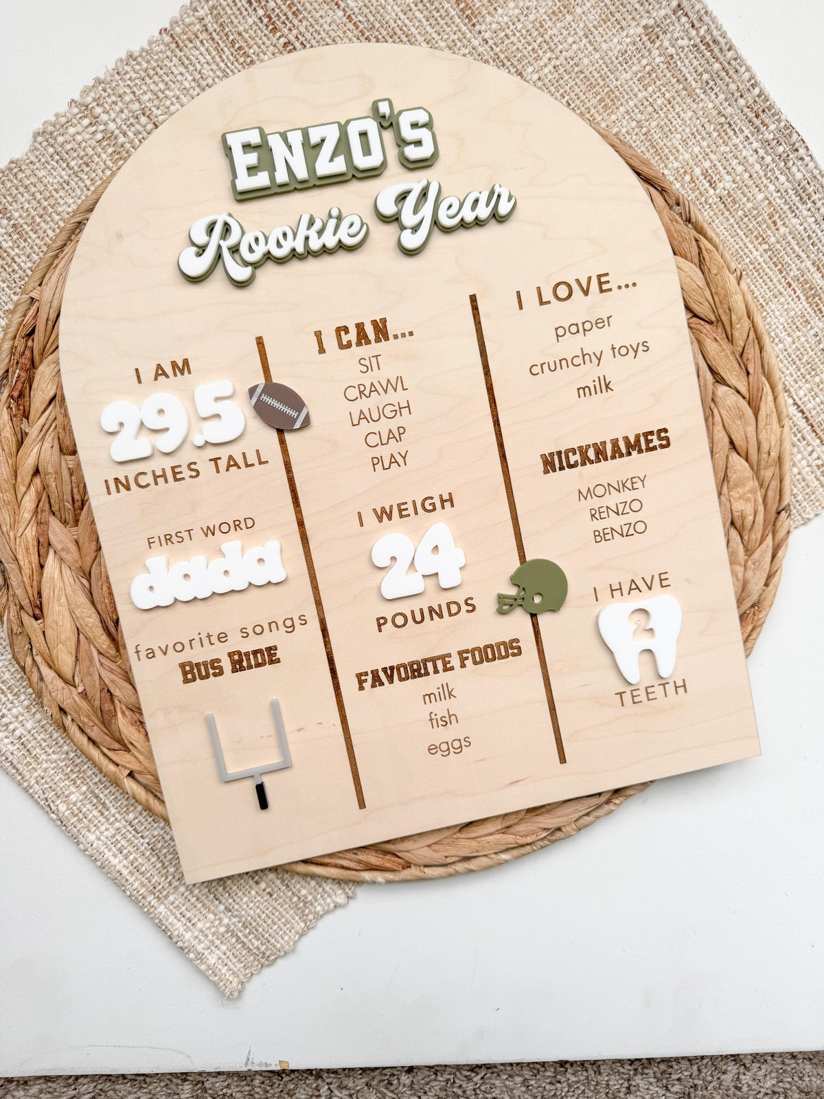 Overhead view of a personalized first birthday stat board for a football party, with a custom name and Rookie Year title in green and white acrylic, engraved milestone sections and white acrylic numbers on an arched maple wood sign resting on a woven placemat