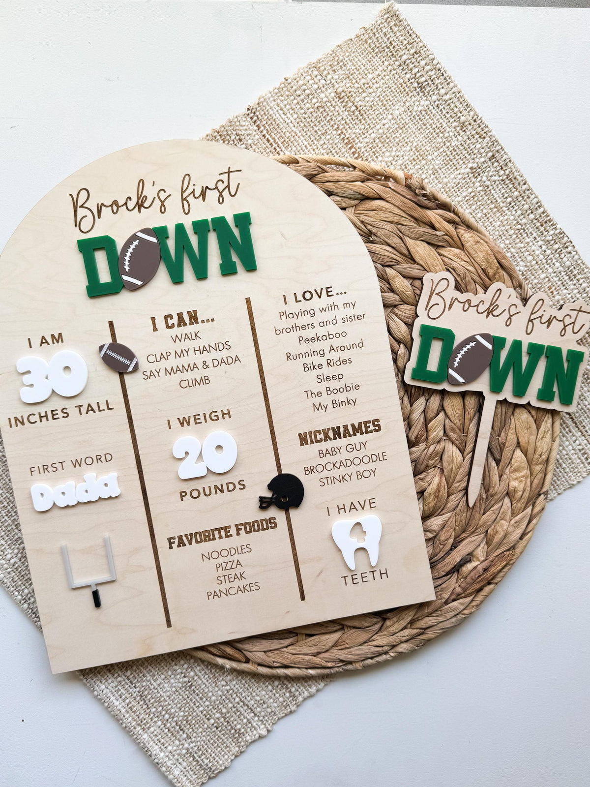 First Down football first birthday milestone board shown with a coordinating personalized First Down acrylic cake topper in matching green, brown and white, a set of football first birthday party decor styled on a woven placemat