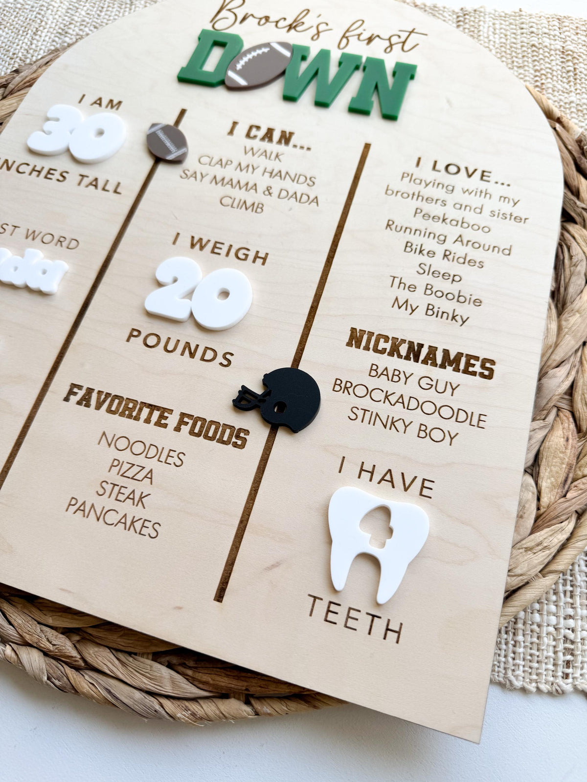 Angled detail of a personalized baby stats board showing engraved I love and nicknames lists, a black acrylic football helmet, engraved favorite foods, white acrylic weight numbers and a white acrylic tooth showing number of teeth on maple wood