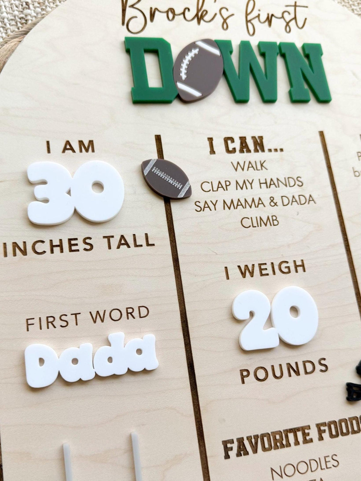 Close-up of a personalized First Down milestone sign showing green acrylic lettering with a football accent, raised white acrylic height and weight numbers, a white acrylic first word and an engraved I can list on natural maple wood