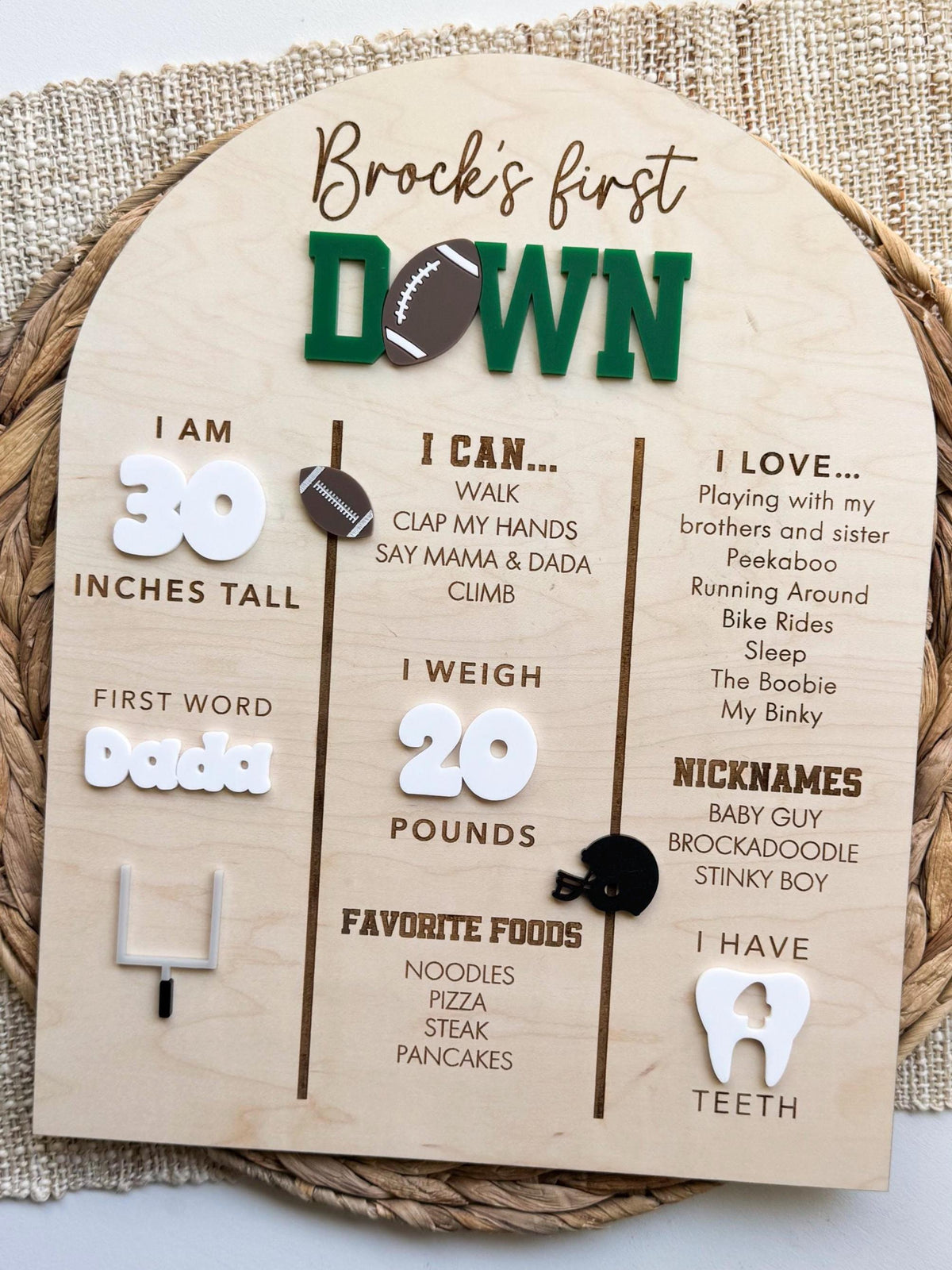 Handmade football birthday sign with an arched maple wood base, three engraved columns of first year milestones, white acrylic numbers, a white acrylic tooth showing number of teeth and football, helmet and goal post accents, displayed on a woven placemat