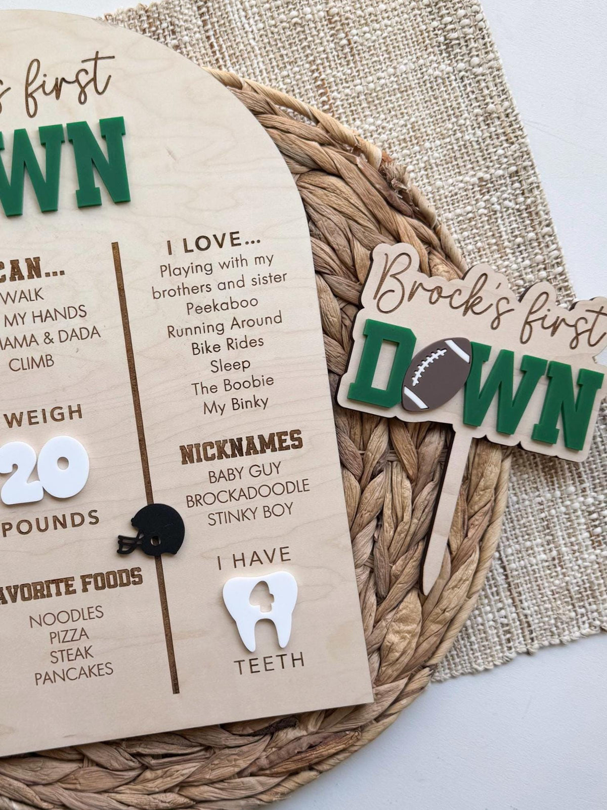 Close view of a matching First Down cake topper with a script name and green acrylic lettering beside the milestone board, showing engraved I love and nicknames lists, a black acrylic helmet and a white acrylic tooth on maple wood
