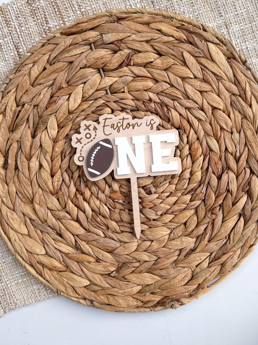 Personalized football first birthday cake topper with child's name, brown layered football, white ONE lettering and football play design on natural maple wood.