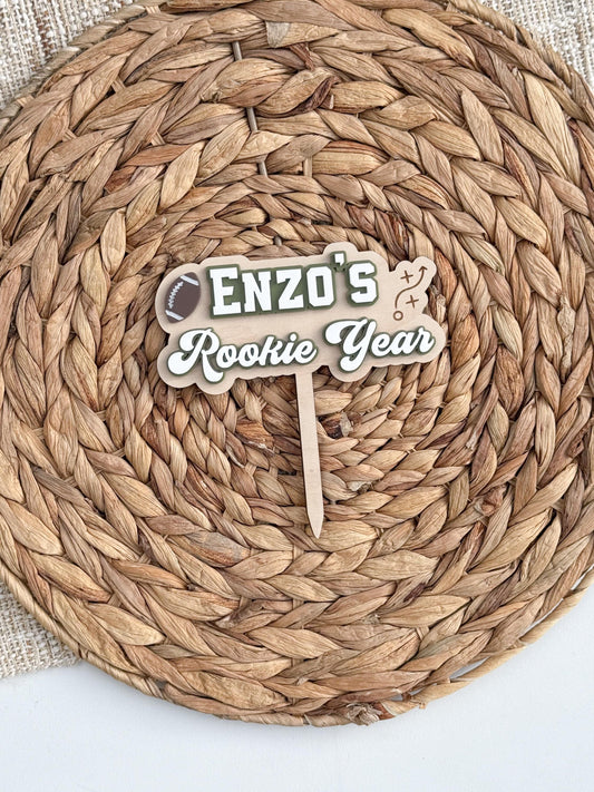 Personalized Rookie Year football first birthday cake topper with child's name, brown football, white and green layered acrylic lettering and natural wood backer.