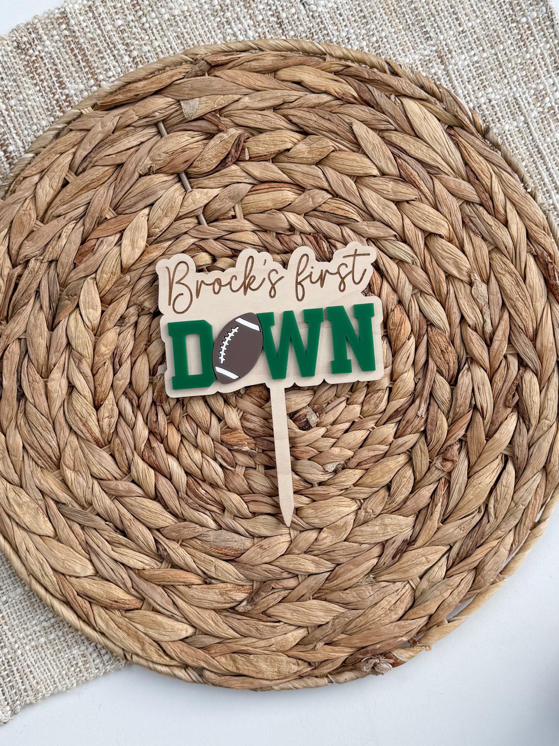 Personalized First Down football cake topper with engraved name on a maple wood backer, green layered acrylic DOWN lettering and a football in place of the O, shown on a woven placemat