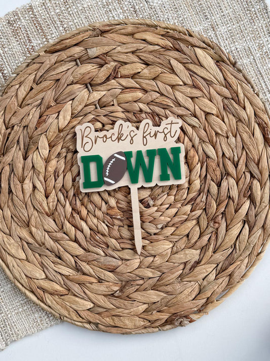 Personalized First Down football cake topper with engraved name on a maple wood backer, green layered acrylic DOWN lettering and a football in place of the O, shown on a woven placemat